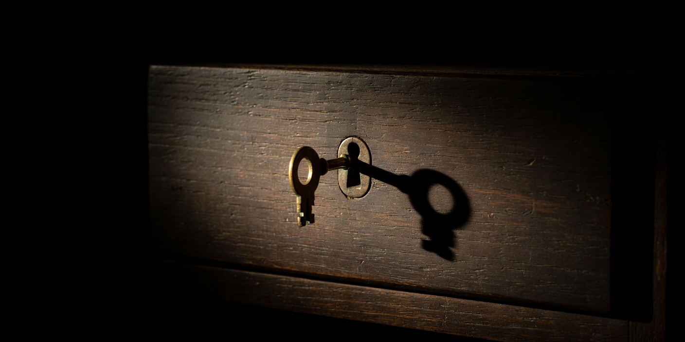 A small brass key in the lock of a wooden drawer, lit from one side in a dark room